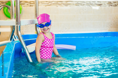 A female child in a pink bathing suit, in a rubber hat and blue glasses sits on the stairs in the pool and laughs and plays with water.