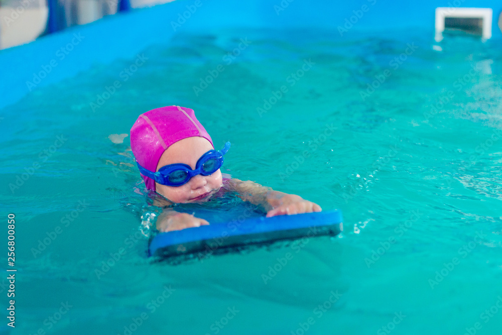 A female child in a pink rubber hat and blue glasses is swimming in a pool with a swimming board.