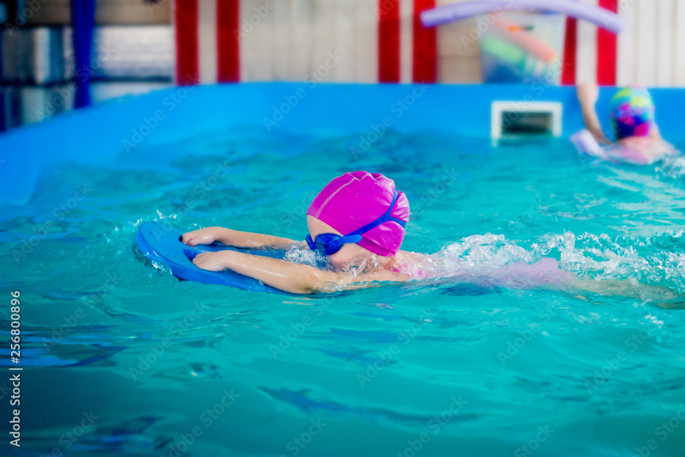 A female child in a pink rubber hat and blue glasses is swimming in a pool with a swimming board.