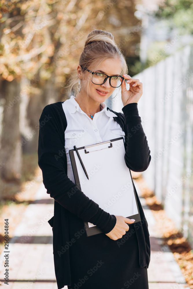 Beautiful trendy blond business woman in glasses with folder of documents in her hands outdoors. Beautiful trendy blond business woman in glasses with folder of documents in her hands outdoors.