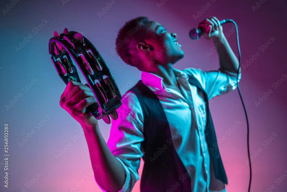 Φωτογραφία African American handsome jazz musician playing tambourine and singing into the microphone in the studio on a neon background