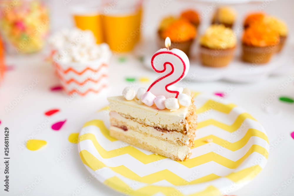 anniversary, celebration and festive concept - piece of cake with candle in shape of number two on plate at birthday party anniversary, celebration and festive concept - piece of cake with candle in shape of number two on plate at birthday party