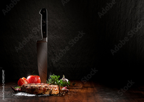 Beef steak on a wood desk with chef knife. Around are fresh tomatoes, sweet and spicy peppers, greens and spices, white salt and black pepper. Creative composition