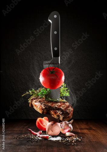 Beef steak on a wood desk with chef knife. Around are fresh tomatoes, sweet and spicy peppers, greens and spices, white salt and black pepper. Creative composition
