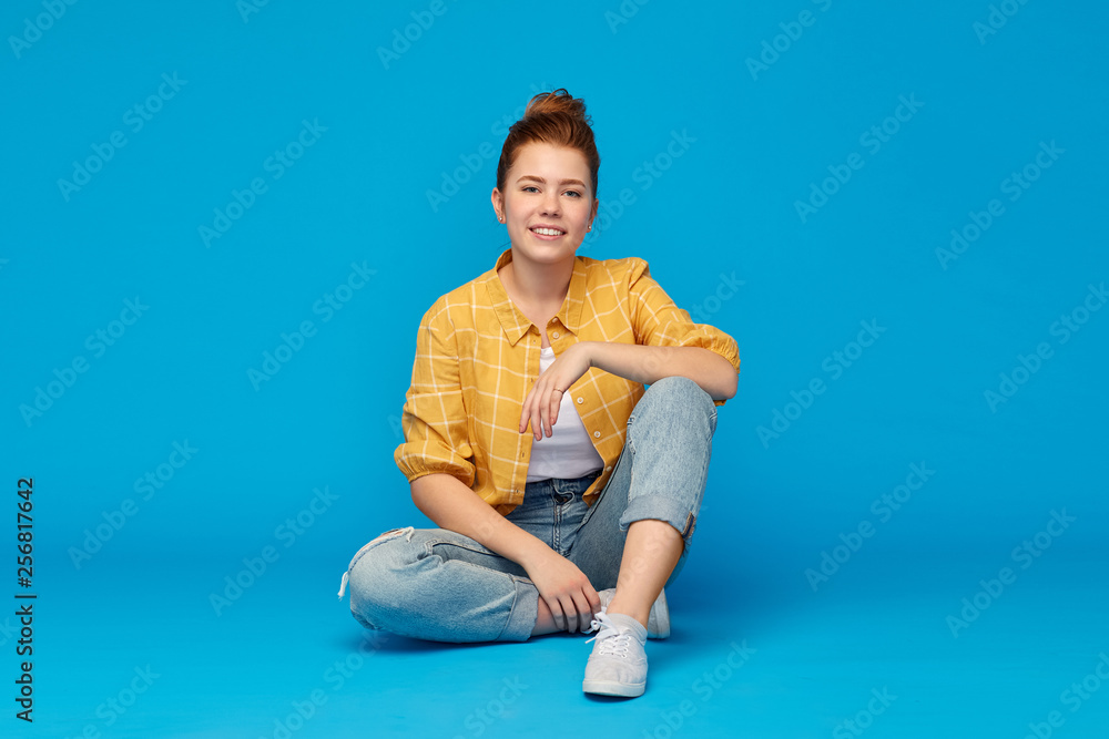 people concept - smiling red haired teenage girl in checkered shirt and torn jeans sitting on floor over bright blue background people concept - smiling red haired teenage girl in checkered shirt and torn jeans sitting on floor over bright blue background