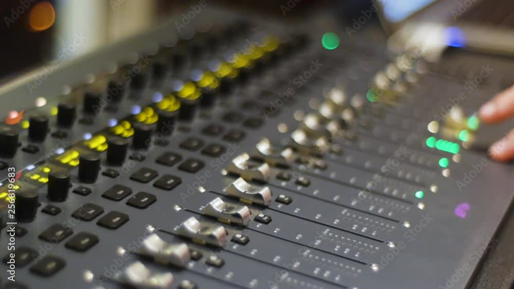Recording studio digital audio mixer faders and sliders being operated by hand w/ depth of field and soft background focus.