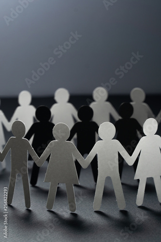 Three human paper figures surrounded by circle of paper people holding hands on dark surface. Bulling, conflict, segregation concept.