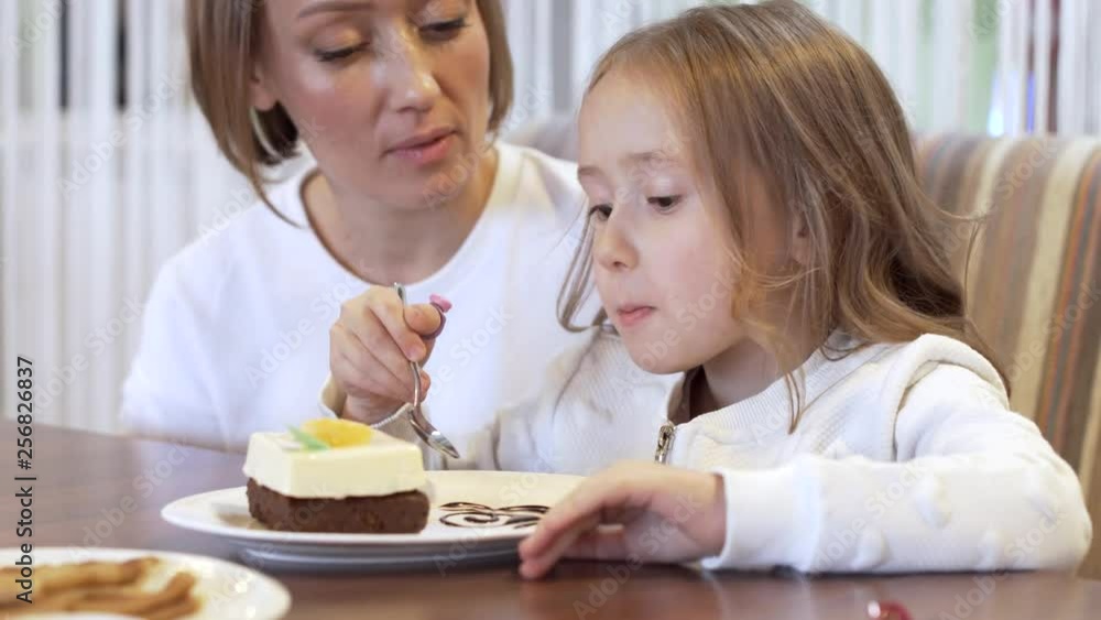 Side View - Charming Girl Eats Dessert Sitting Next To Beautiful Young Mother Side View - Charming Girl Eats Dessert Sitting Next To Beautiful Young Mother