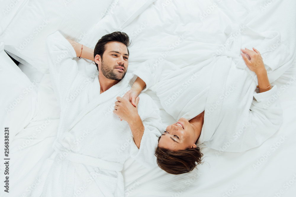 top view of happy couple in white bathrobes holding hands while lying on bed top view of happy couple in white bathrobes holding hands while lying on bed