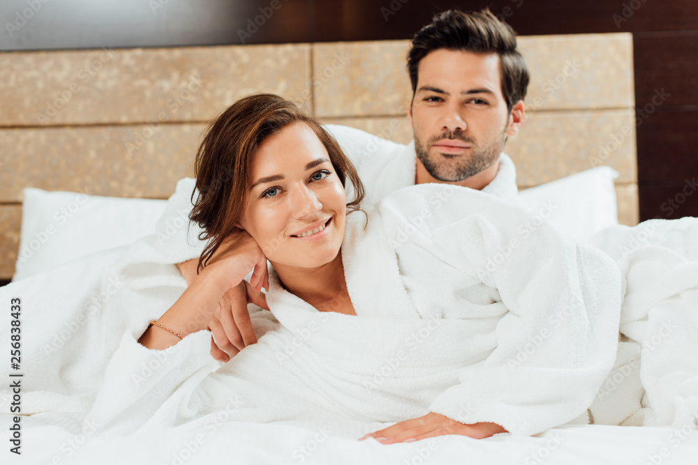 Fototapeta premium handsome man and happy girlfriend in white bathrobes looking at camera while lying on bed
