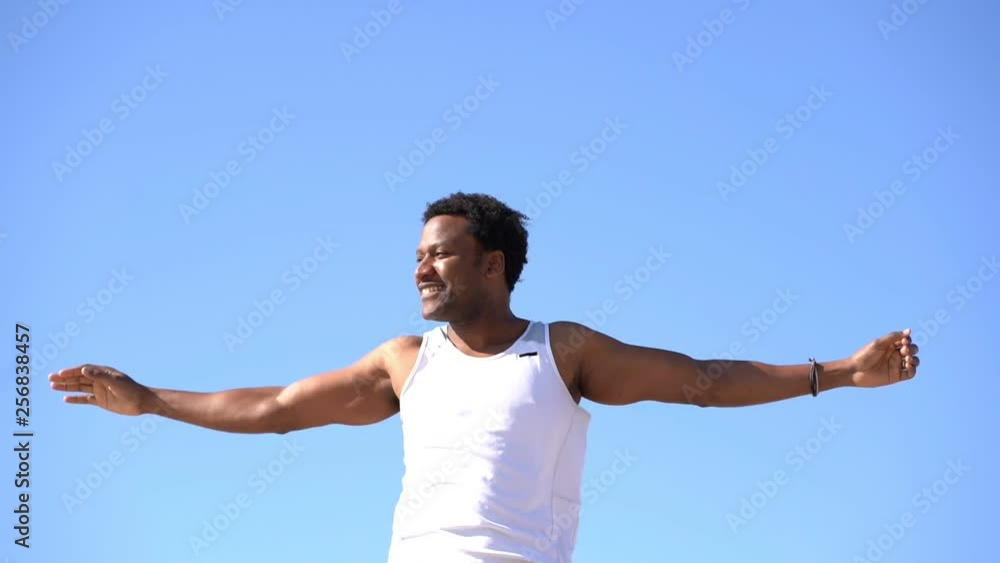 Smiling young man exercising against blue sky. Low angle view of happy african american sportsman stretching arms and looking away at sunny windy day, handheld shot. Workout concept Smiling young man exercising against blue sky. Low angle view of happy african american sportsman stretching arms and looking away at sunny windy day, handheld shot. Workout concept