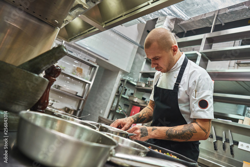 Inspiration in cooking. Young male chef with several tattoos on his arms is garnishing italian pasta in a restaurant kitchen.