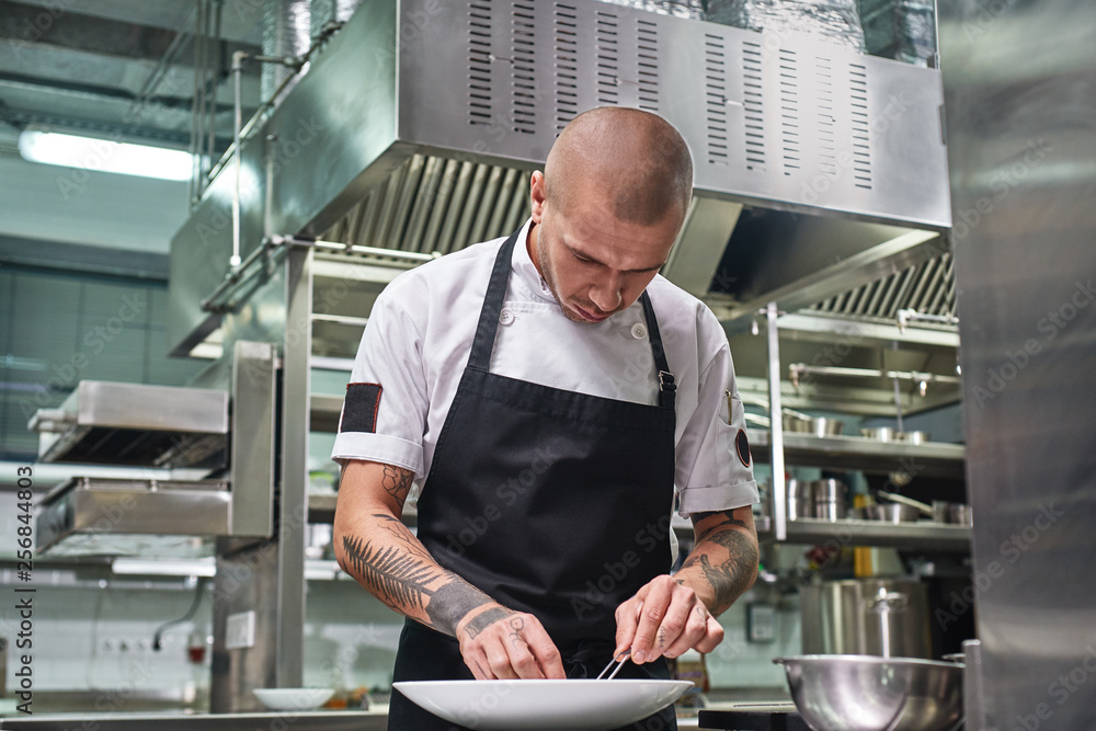 Finishing a dish. Attractive male chef with beautiful tattoos on his arms garnishing his dish on the plate in restaurant kitchen