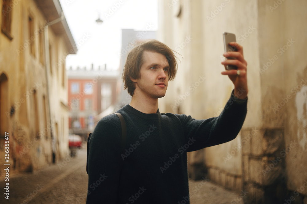 A man travels in Europe street. A man smiles, walks through the streets of the old city, with a briefcase. Student travels alone. A young man takes pictures on the camera phone. A man taking pictures