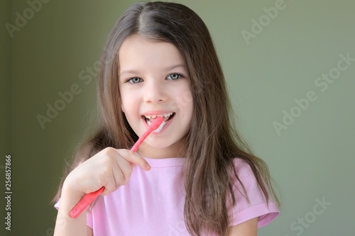 Caucasian beautiful girl brushing her teeth. Long haired smiling white girl brushing teeth with pink toothbrush on green background. Green eyed cute child in pink tshirt doing her morning hygiene. 