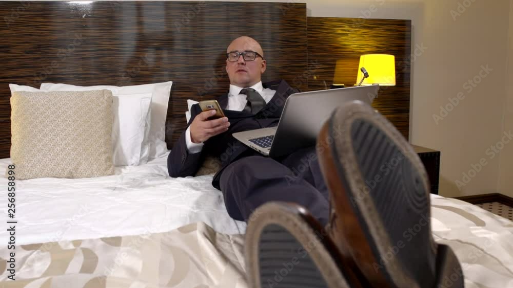 tired businessman is relaxing in bedroom of hotel in evening, he is wearing suit, typing on phone tired businessman is relaxing in bedroom of hotel in evening, he is wearing suit, typing on phone
