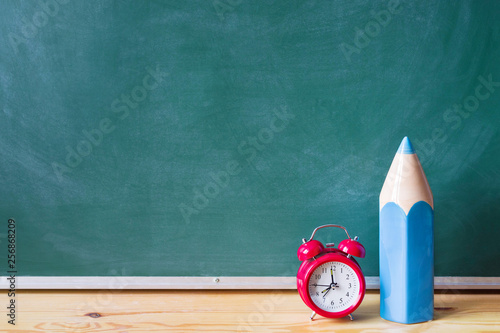 Back to school. Blue pencil and red alarm clock on a wooden surface against a green blackboard. School background with space for your text