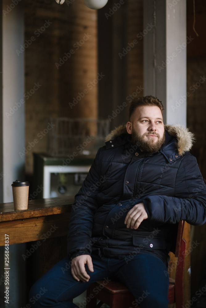 Fototapeta premium A man with a beard in a warm winter jacket poses for advertising men's clothing in the winter. Advertising of men's clothing