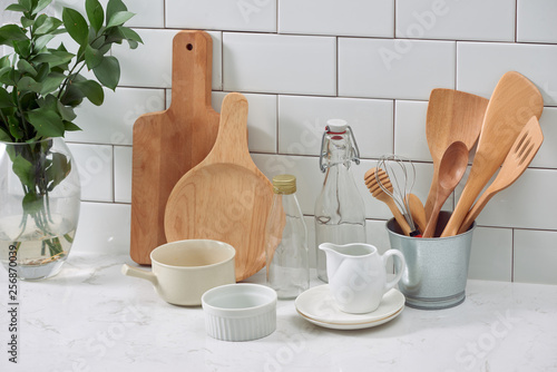 Simple rustic kitchenware against white wooden wall  rough ceramic pot with wooden cooking utensil set  stacks of ceramic bowls  jug and wooden trays.