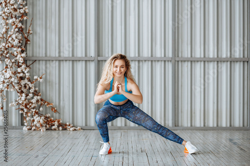 fitness and sports concept. A pretty blonde girl in a blue sports uniform is training in the hall. Woman doing lunges or stretching and looking at the camera smiling