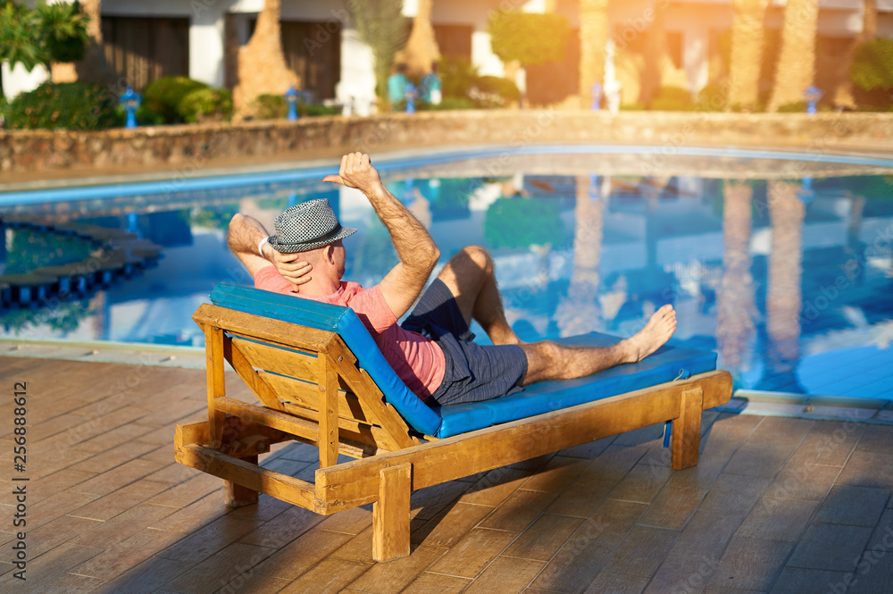 Relax in the pool summer. Young and successful man in hat lying on a sun lounger at the hotel on the background of sunset, concept time to travel