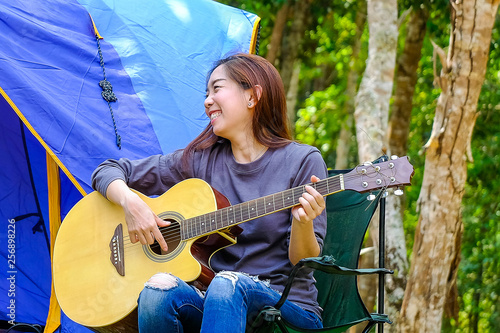 Curly blond long hair asian teenager girl with long shrit and white shorts, playing guitar front of blue canvas camping tent on grass field with smile happy face photo