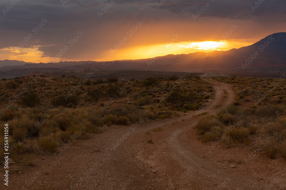 Sunset light glows orange over a landscape with a dirt road leading into the desert with the sun going down behind clouds and a distant mountain.