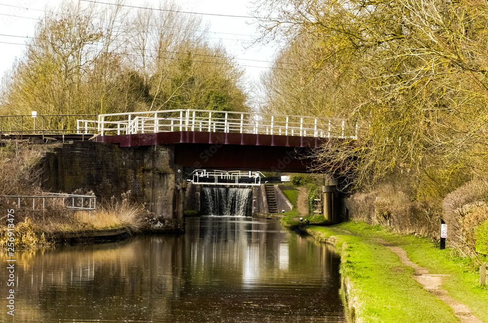 The London-Rugby railway bridge crosses over the Grand Union Canal in Whilton Locks. Landscape looking up the waterway, with water cascading through the closed lock gates. Northamptonshire. England. The London-Rugby railway bridge crosses over the Grand Union Canal in Whilton Locks. Landscape looking up the waterway, with water cascading through the closed lock gates. Northamptonshire. England.
