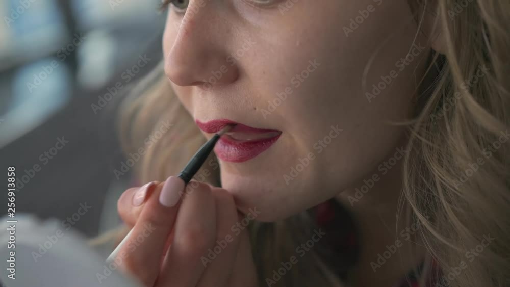 Slow Motion A young beautiful unrecognized girl applies lipstick to her lips with a brush and looks at herself in a small mirror sitting in the departure area of the international airport terminal Slow Motion A young beautiful unrecognized girl applies lipstick to her lips with a brush and looks at herself in a small mirror sitting in the departure area of the international airport terminal
