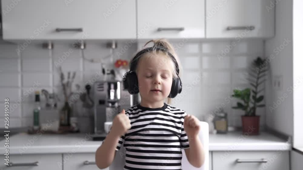 Little girl in a striped T-shirt sits on a chair listening to music in headphones, sings,dance and smiles 4k Little girl in a striped T-shirt sits on a chair listening to music in headphones, sings,dance and smiles 4k