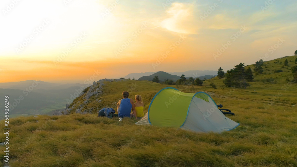 AERIAL: Active young couple sitting by their tent and watching the sunset.