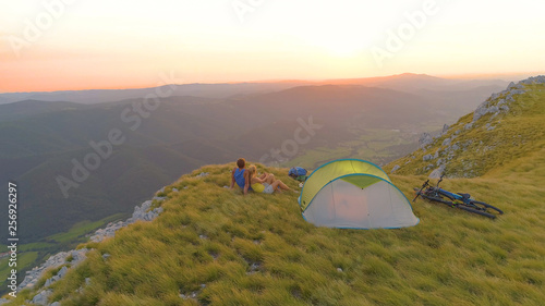 AERIAL  Flying over mountain biking couple cuddling by their tent at sunset.