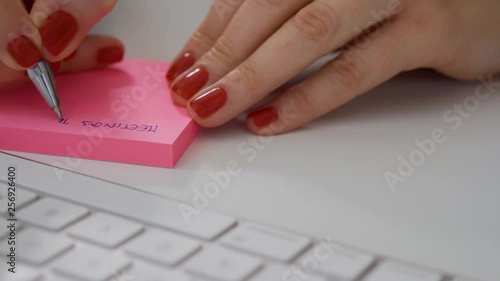 Close up of female hands with red nails write note on pink post it next to the white computer keyboard