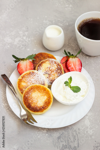 Cottage cheese fritters (syrniki) served with sour cream, fresh berries (strawberry and blueberry) and mint on a light grey background. Healthy breakfast or diet lunch. Copy space.