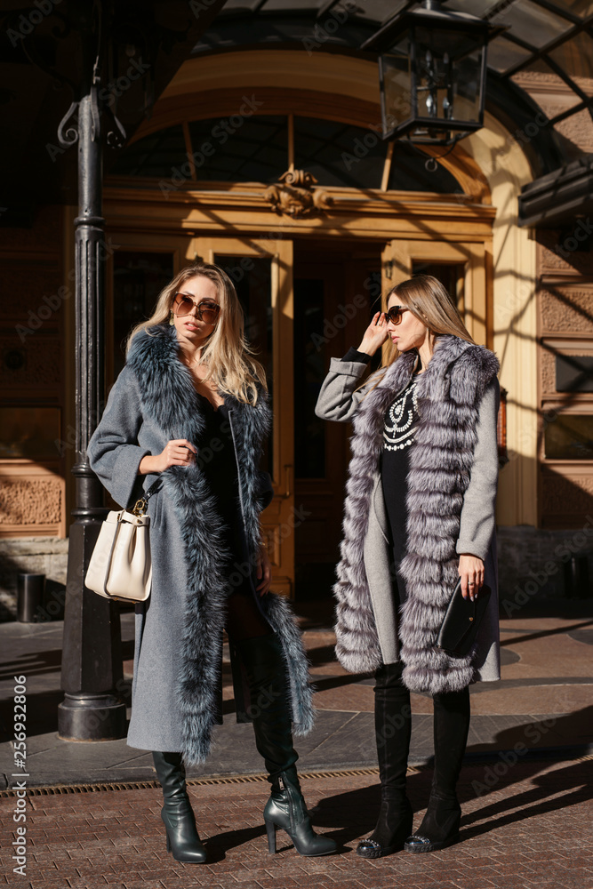 Two beautiful women in sun glasses and fur coat, smiling, posing with luxury bags and accesories while standing and walking outdoors. Female fashion city lifestyle spring or autumn shopping concept