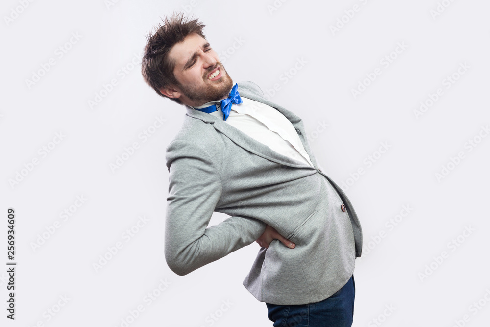 spine or kidney pain. Profile side view portrait of young bearded businessman in casual grey suit and blue bow tie standing and holding his painful back. studio shot, isolated on light grey background spine or kidney pain. Profile side view portrait of young bearded businessman in casual grey suit and blue bow tie standing and holding his painful back. studio shot, isolated on light grey background