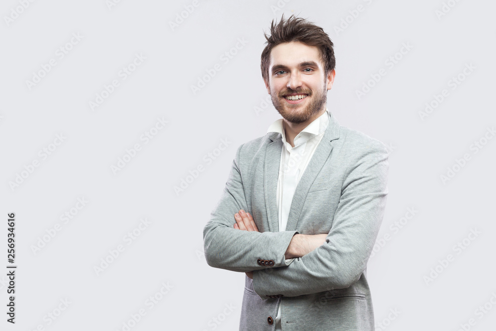 Portrait of happy satisfied handsome bearded man in white shirt, casual grey suit standing, crossed hands and looking at camera with toothy smile. indoor studio shot, isolated on light grey background Portrait of happy satisfied handsome bearded man in white shirt, casual grey suit standing, crossed hands and looking at camera with toothy smile. indoor studio shot, isolated on light grey background