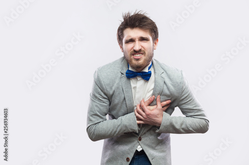 hearth pain or attack. Portrait of worried handsome bearded man in casual grey suit and blue bow tie standing and holding his painful heart. indoor studio shot, isolated on light grey background.