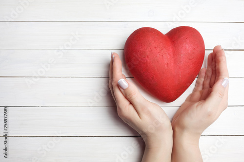 Woman holding hands near red heart on white wooden background, top view