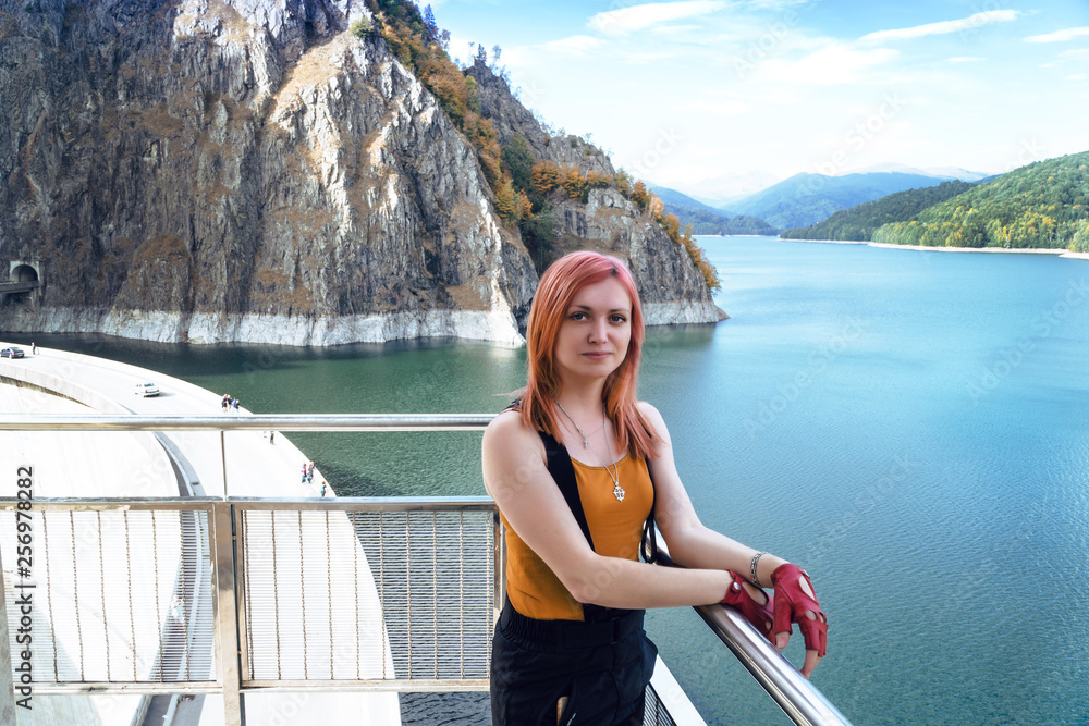 girl traveler with red hair posing in background Dam lake Vidraru in Romania, autumn sunny day