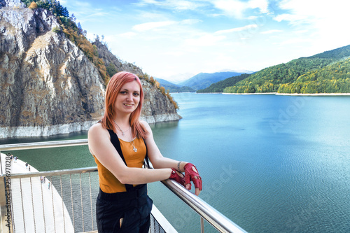 girl traveler with red hair posing in background Dam lake Vidraru in Romania, autumn sunny day