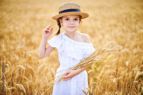 Happy child in autumn wheat field. Beautiful girl in white dress and straw hat have fun with playing, harvesting wheat.