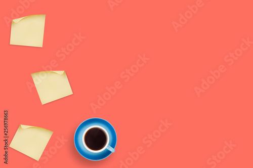 Workplace on table of coral color with one blue ceramic cup full of black coffee on saucer and blank square paper stickers with curved corner. Top view. Office work concept. Copy space