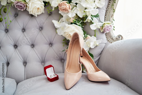 Beautiful beige bride shoes on high heels and earings on lavender chesterfield chair with flowers, copy space. Luxury bridal accessories and jewelry. Wedding morning preparations photo