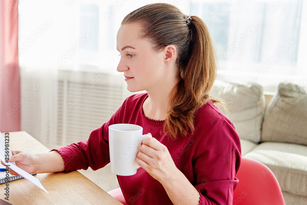 Portrait of young woman sitting at wood desk in office with cup of coffee and reading documents, copy space. Businesswoman doing his work at home Portrait of young woman sitting at wood desk in office with cup of coffee and reading documents, copy space. Businesswoman doing his work at home
