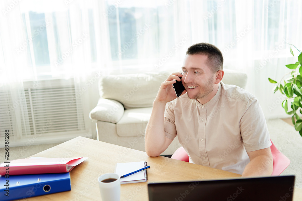 Happy young businessman sitting at wood desk, working on laptop and talking on mobile phone with client at home office, copy space Happy young businessman sitting at wood desk, working on laptop and talking on mobile phone with client at home office, copy space