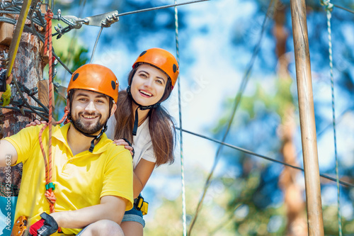 Young woman and man in protective gear are sitting on rope bridge hanging on high trees, posing and smiling. Rope adventure park with obstacles and ziplines. Extreme rest and summer activities concept