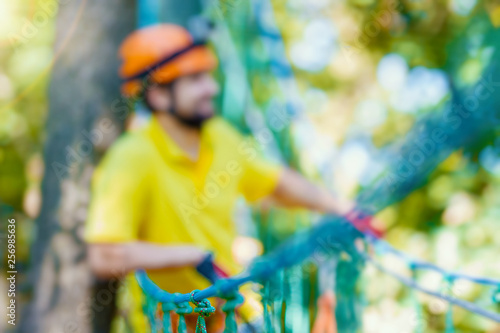 Young male man adult wears protective helmet with action camera having fun in extreme rope park, amusement park. Climbing in rope bridge at green forest. Active healthy lifestyle in summer. Bokeh