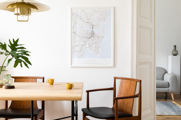 Stylish and eclectic dining room interior with mock up poster map, sharing table design chairs, gold pedant lamp and elegant sofa in second space. White walls, wooden parquet. Tropical leafs in vase.