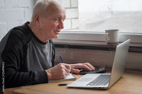 An old man looks at a laptop screen and takes notes in a notebook, selective focus.